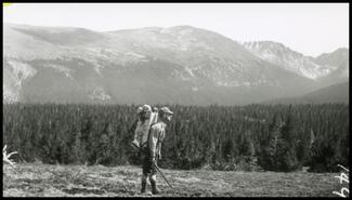 [Man hiking with backpack and rifle, with view of forest and mountains]