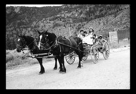 Two horses pulling a democrat wagon with five Interior Salish people