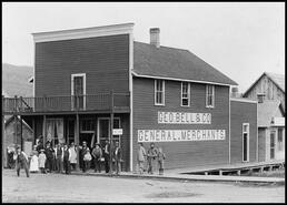 Group watching a man throw a shotput in front of Geo Bell & Co. General Merchants store in Enderby