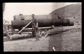 Two men with a log on a truck bed and steam engine boiler in the background at Munson and Simpson Sawmill, Beaver Lake Road